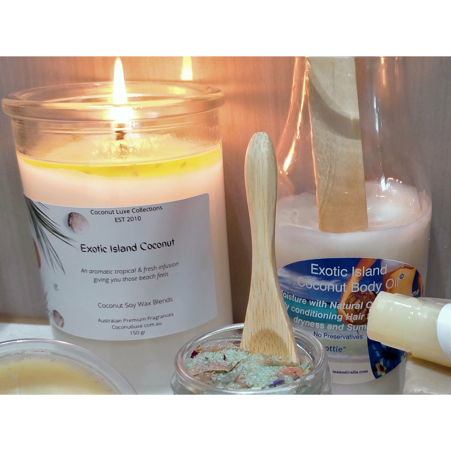 set of exotic island coconut products including a lit soy wax candle, body oil bottle, and a jar with a wooden spoon, creating a tropical and soothing ambiance